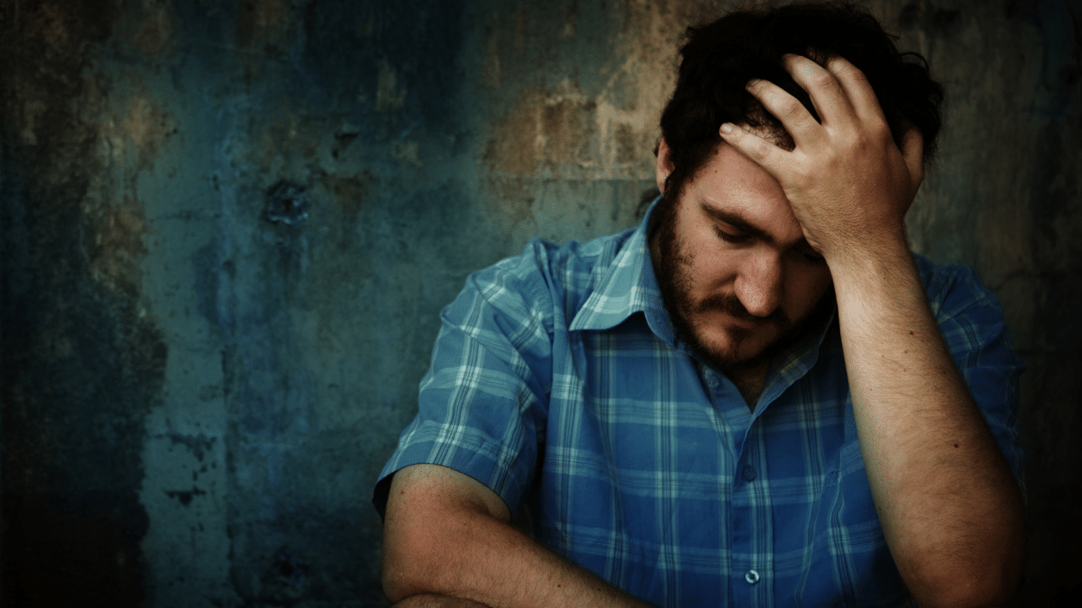 Imagem de destaque do post Porque Deus permite o sofrimento, mostra um Homem de cabeça baixa e com a mão esquerda na cabeça, ele esta de camisa azul xadrez encostado numa parede descascada pelo tempo.